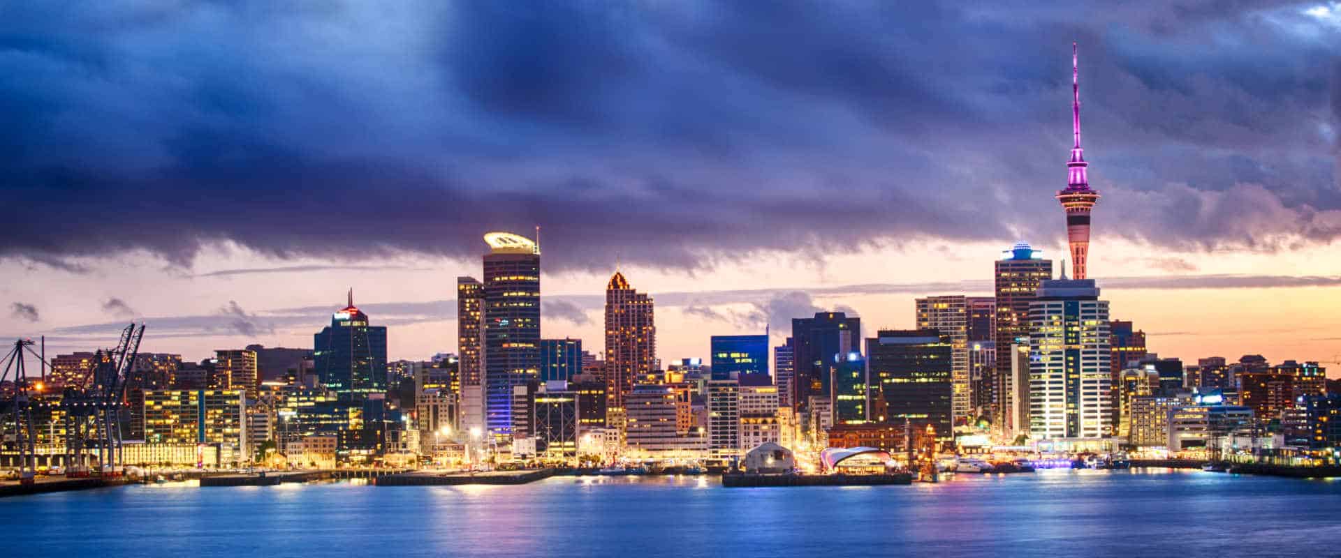 The Auckland city skyline and waterfront at dusk, with the Sky Tower lit above the harbour